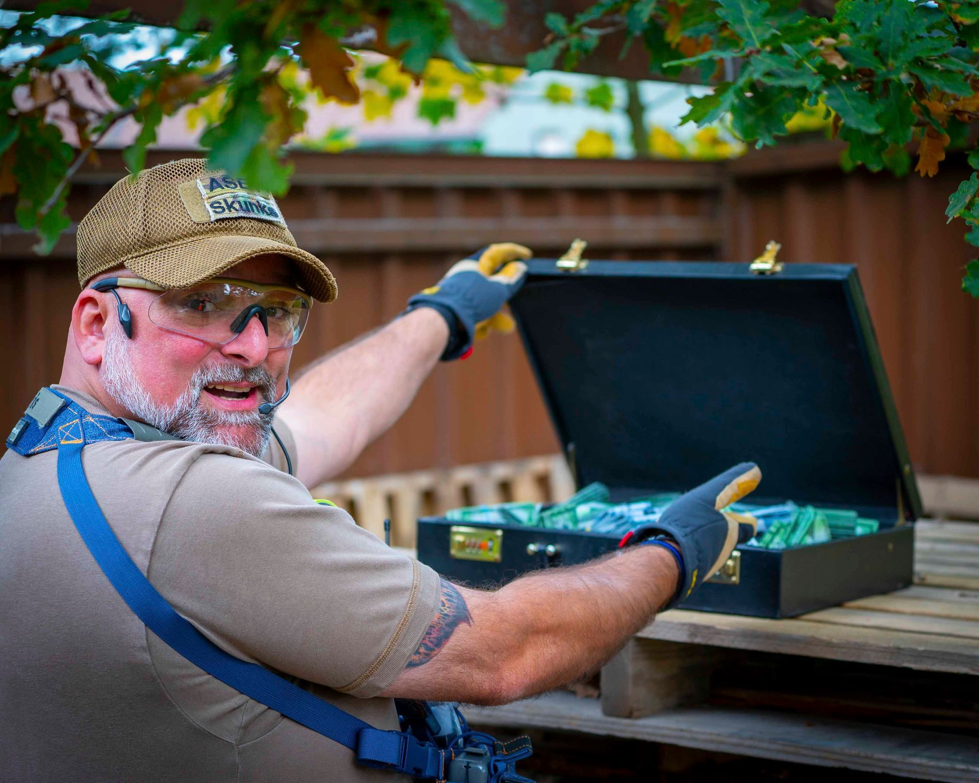 Ein Mann öffnet im Freien einen mit Geld gefüllten Aktenkoffer. Er trägt Mütze, Handschuhe und Schutzbrille und lächelt.