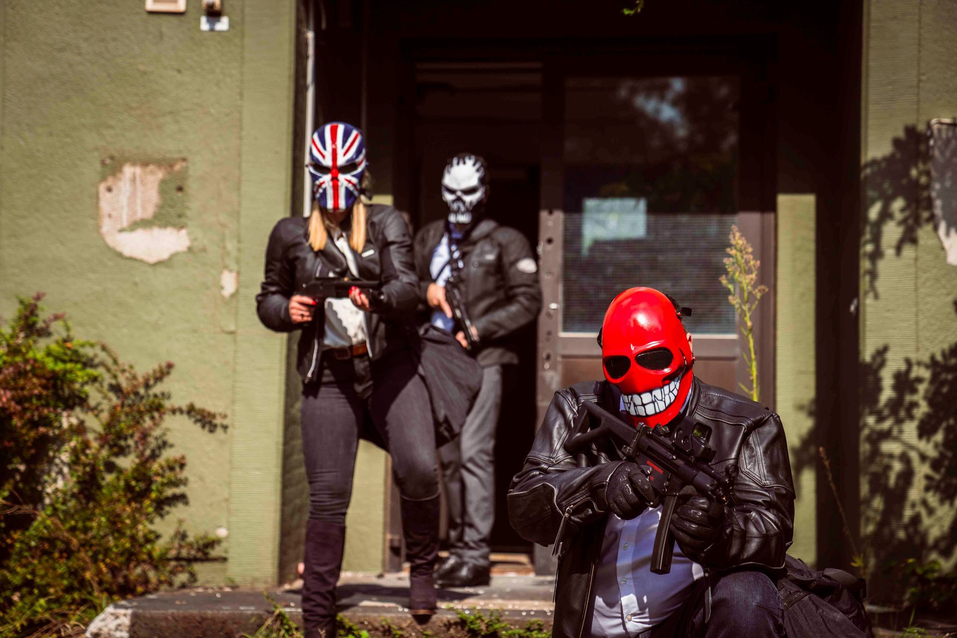 Drei Personen mit Masken und Lederjacken kommen aus einem Gebäude; sie halten Schusswaffen.