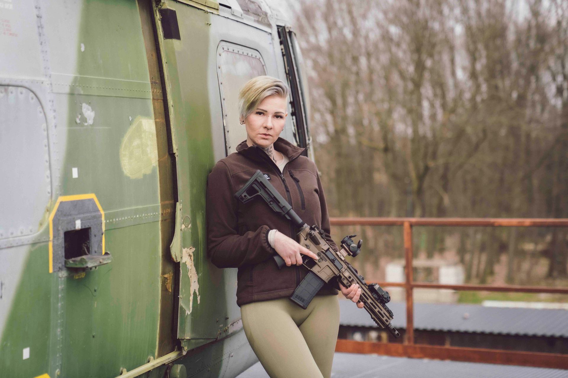 Frau in brauner Jacke und Leggings, die ein Gewehr hält, lehnt an einem grünen Hubschrauber.