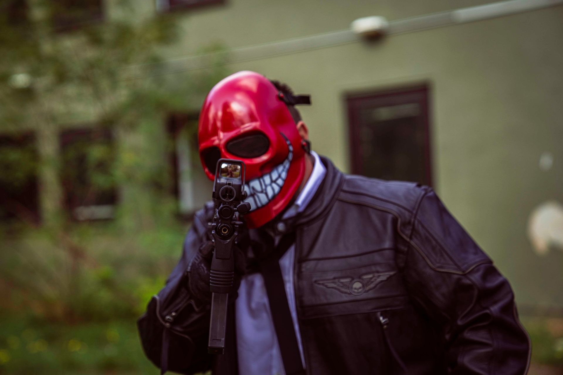 Eine Person mit roter Totenkopfmaske und schwarzer Lederjacke zielt mit einer Pistole.