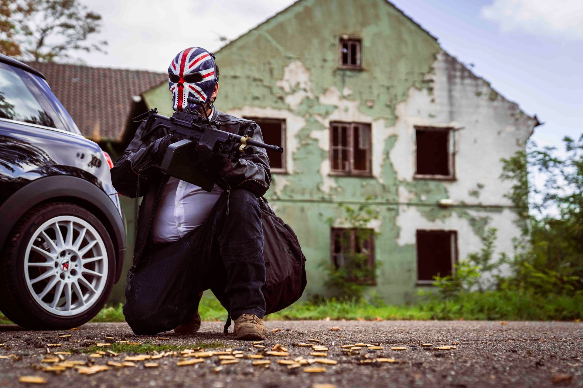 Eine Person mit Union-Jack-Maske kniet mit einem Gewehr neben einem schwarzen Auto