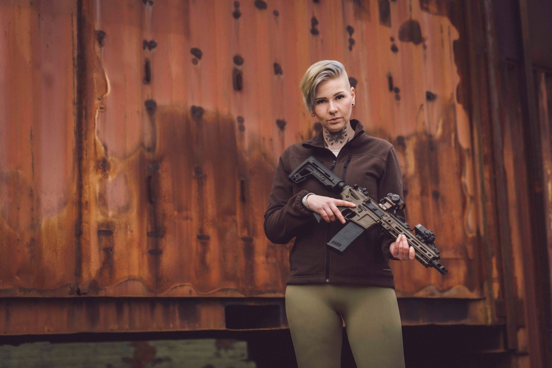 Eine Frau mit kurzen Haaren hält ein Gewehr und steht vor einer rostigen Wellblechwand.