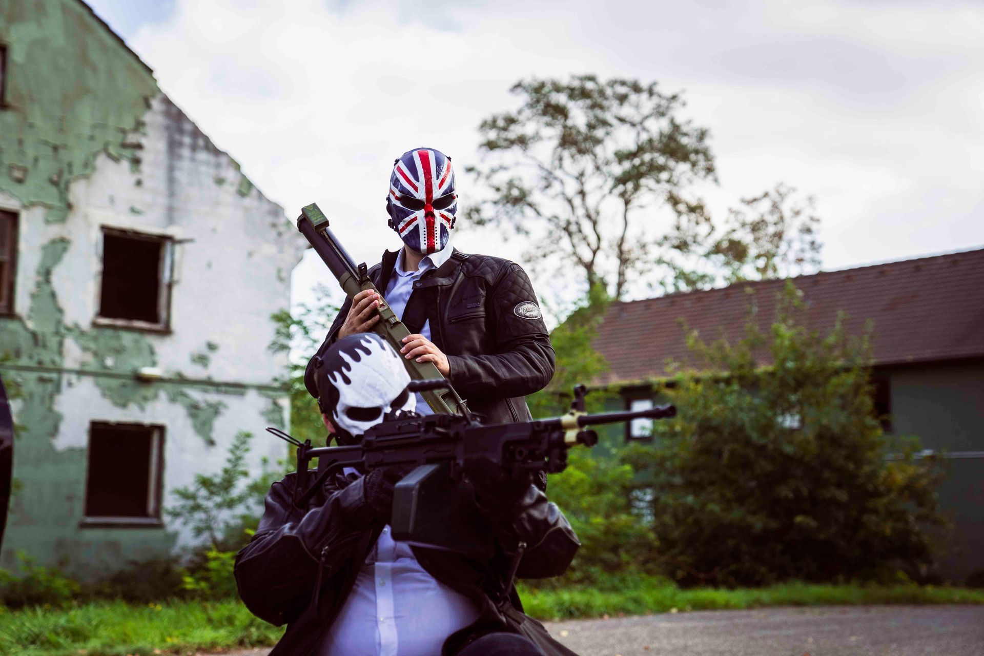 Eine Person mit Union-Jack-Maske hält vor einem verwitterten Gebäude eine Gitarre und ein Maschinengewehr.