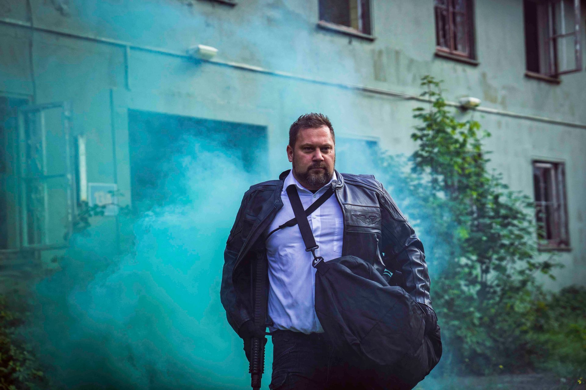 Mann mit Bart in Lederjacke verlässt ein Gebäude, aus dem blauer Rauch aufsteigt. Schwarze Tasche über der Schulter