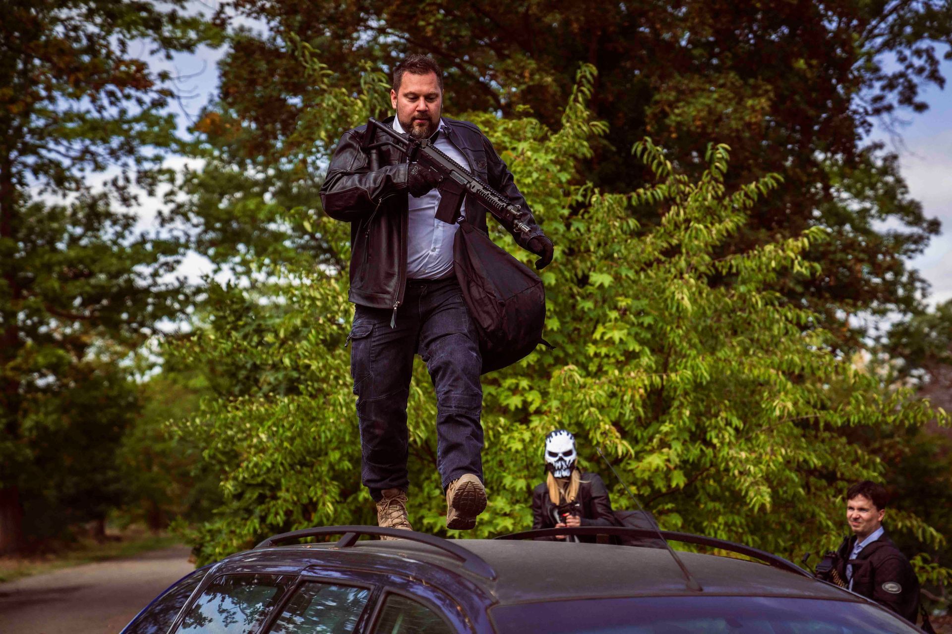 Ein Mann mit einer Pistole springt auf dem Dach eines Autos; im Hintergrund ist eine maskierte Gestalt und ein Mann zu sehen