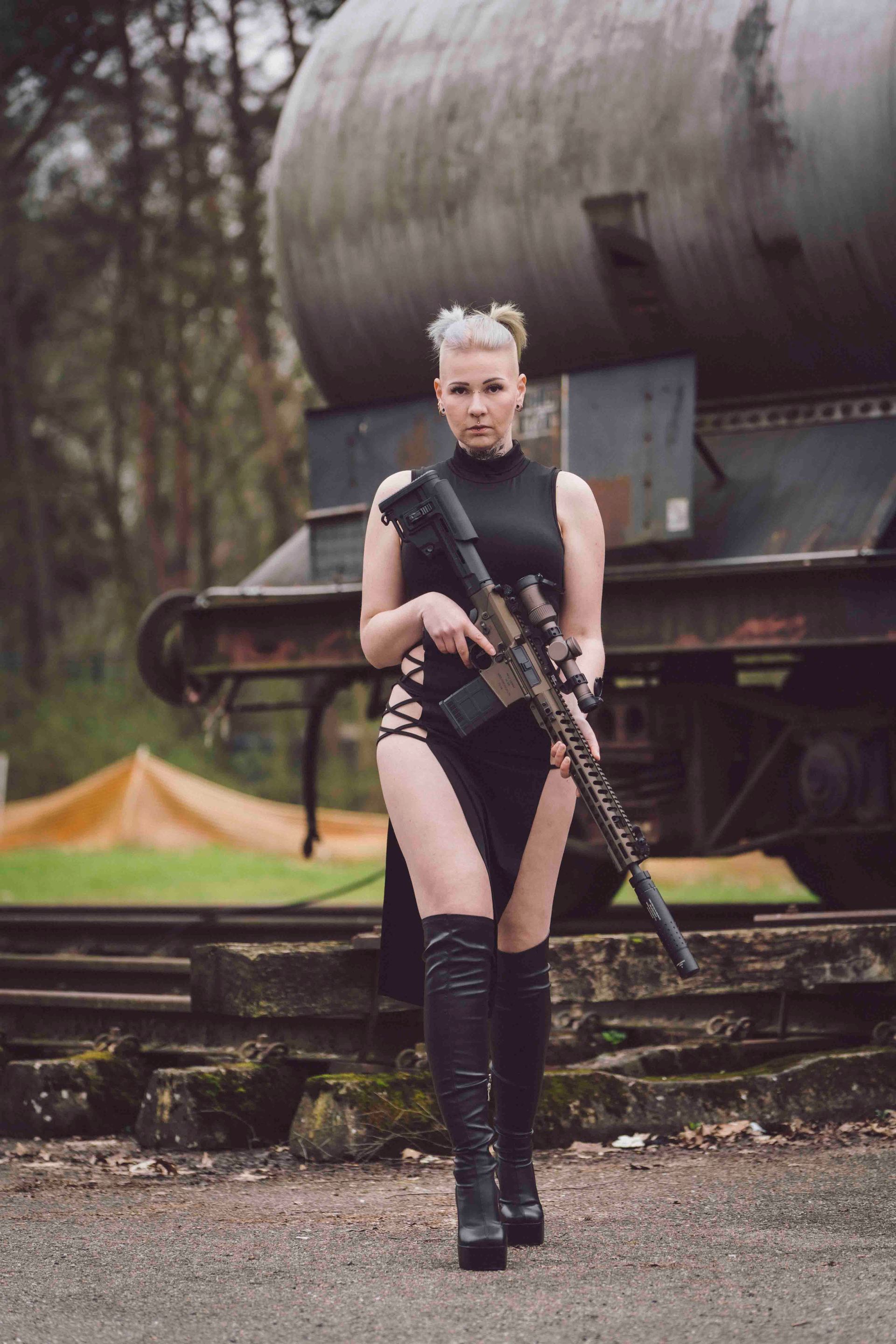 Frau in schwarzem Kleid und Stiefeln hält ein Gewehr und steht in der Nähe von Bahngleisen.