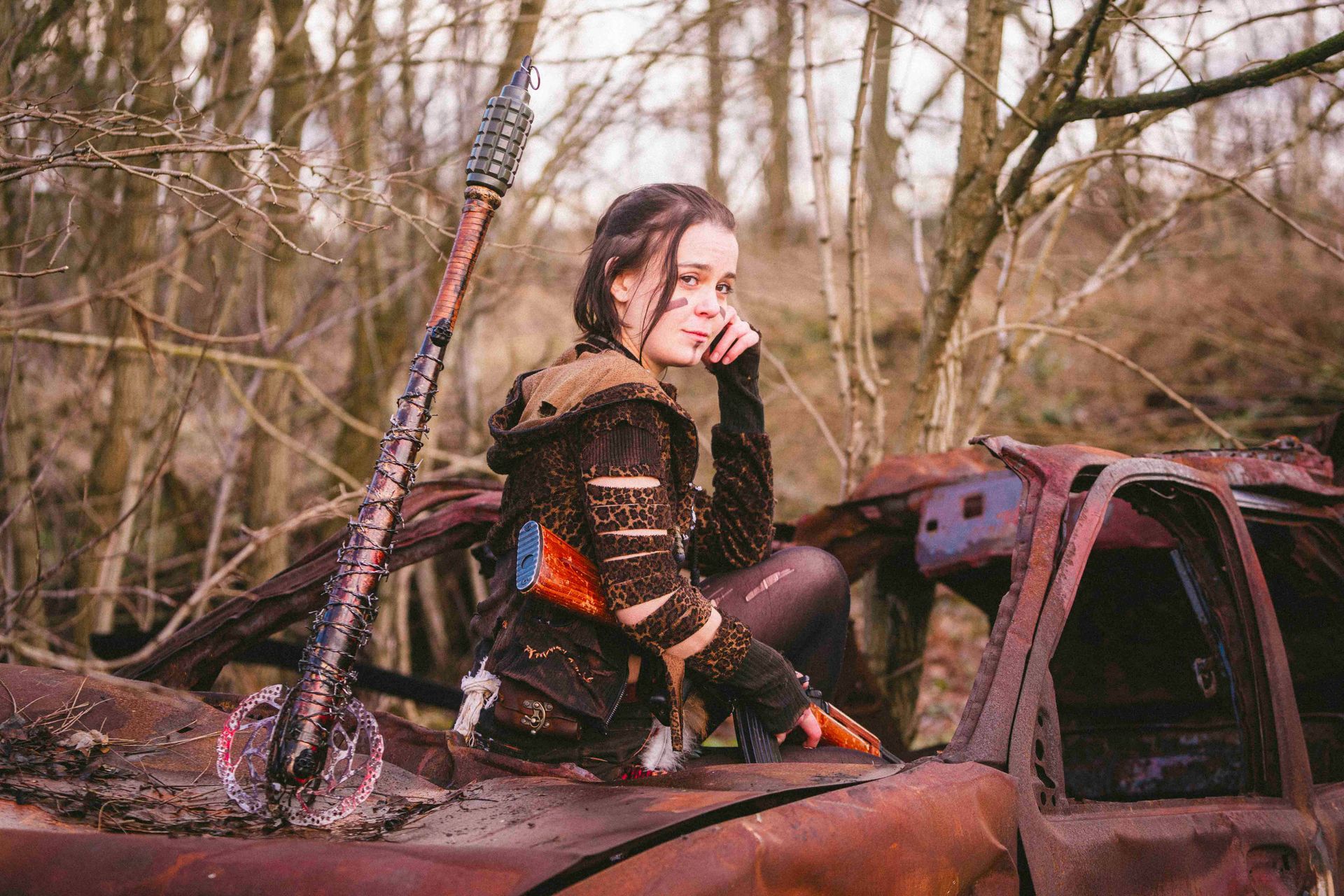 Eine Frau in postapokalyptischer Kleidung lehnt in einer Waldlandschaft an einem verrosteten Auto und hält eine Keule.