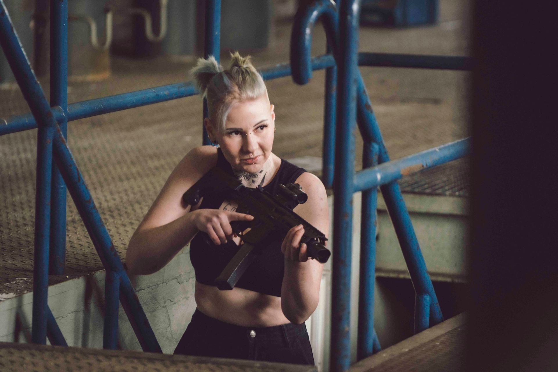 Frau mit Gewehr in der Hand, posiert in einer Industrieumgebung, den Blick zur Seite gerichtet.