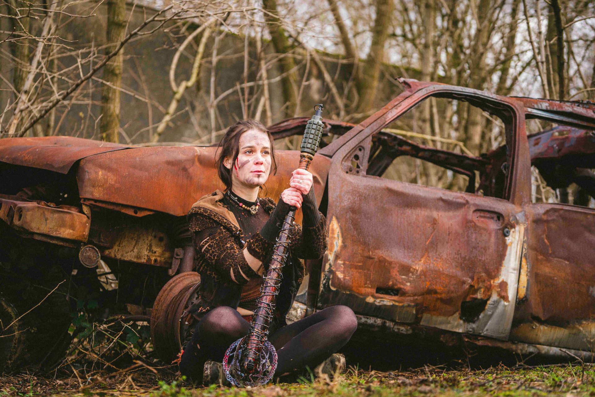 Eine Frau in postapokalyptischer Kleidung sitzt im Schneidersitz vor einem rostigen Autowrack und hält eine Stachelwaffe