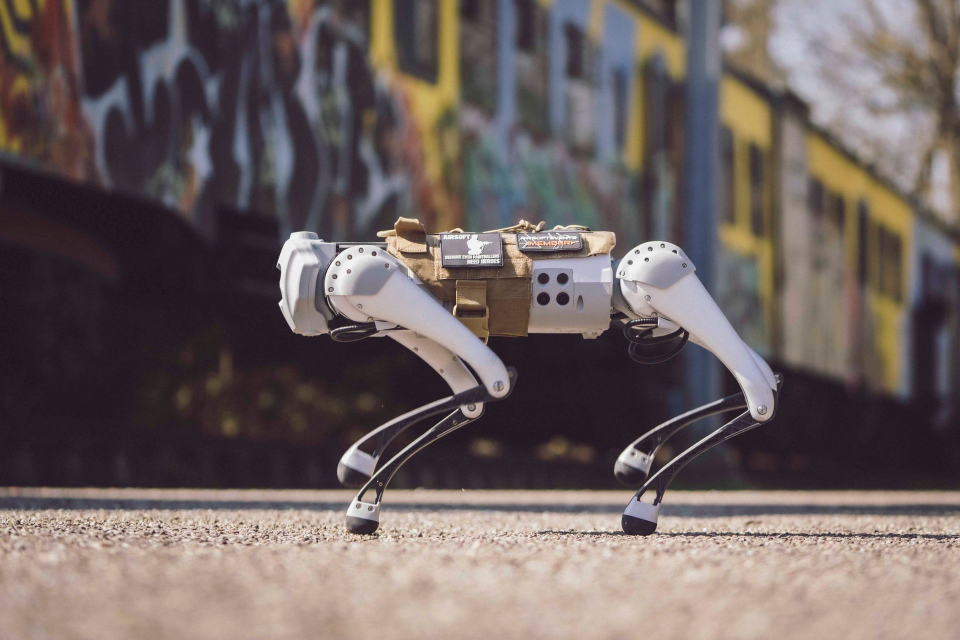 Ein vierbeiniger Roboter auf gepflasterter Fläche bewegt sich vorwärts; im Hintergrund ist ein Gebäude mit Graffiti zu sehen.