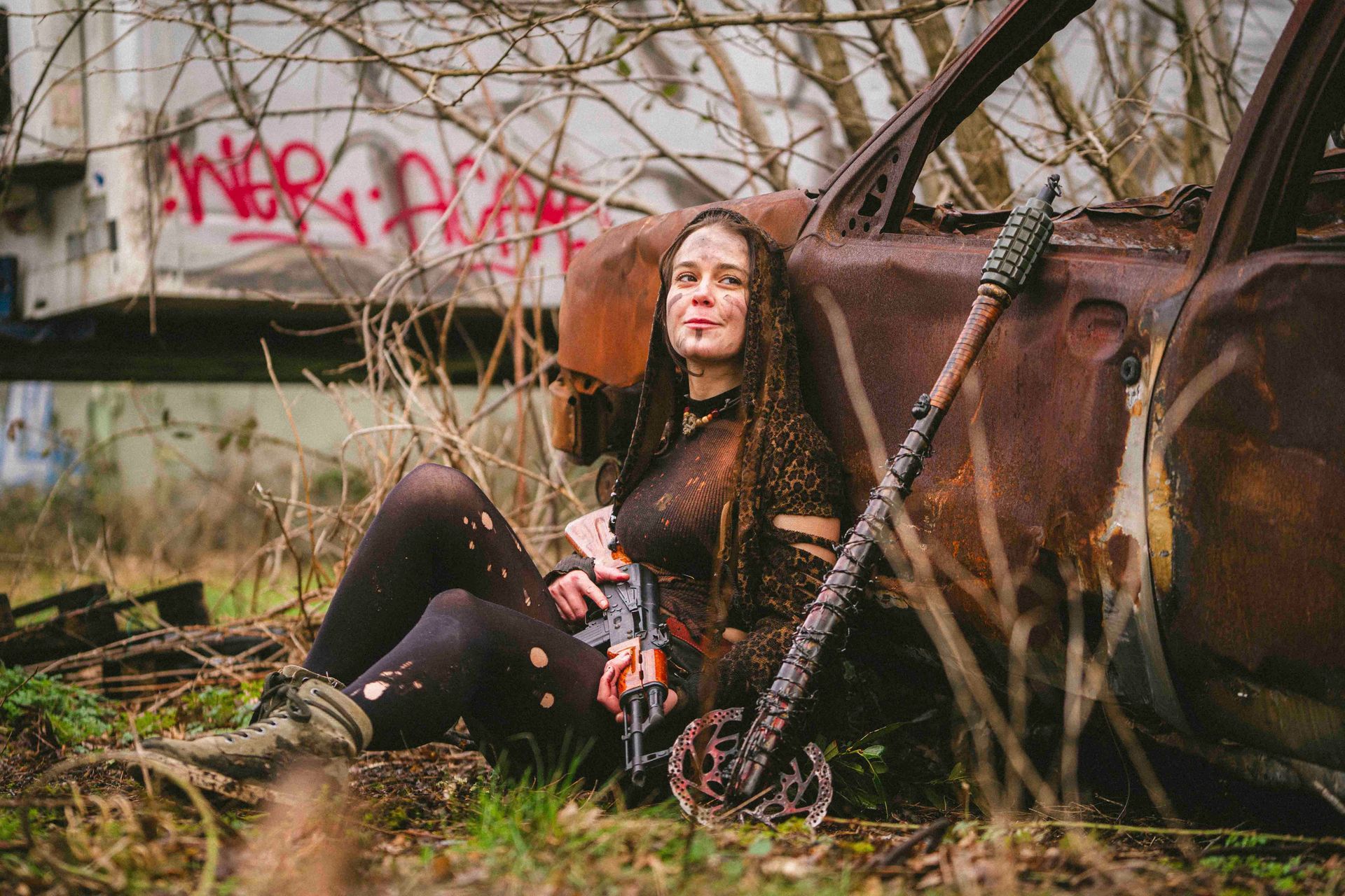 Eine Frau in zerrissener Kleidung mit einer Waffe lehnt an einem verrosteten Auto, umgeben von Graffiti und Trümmern.