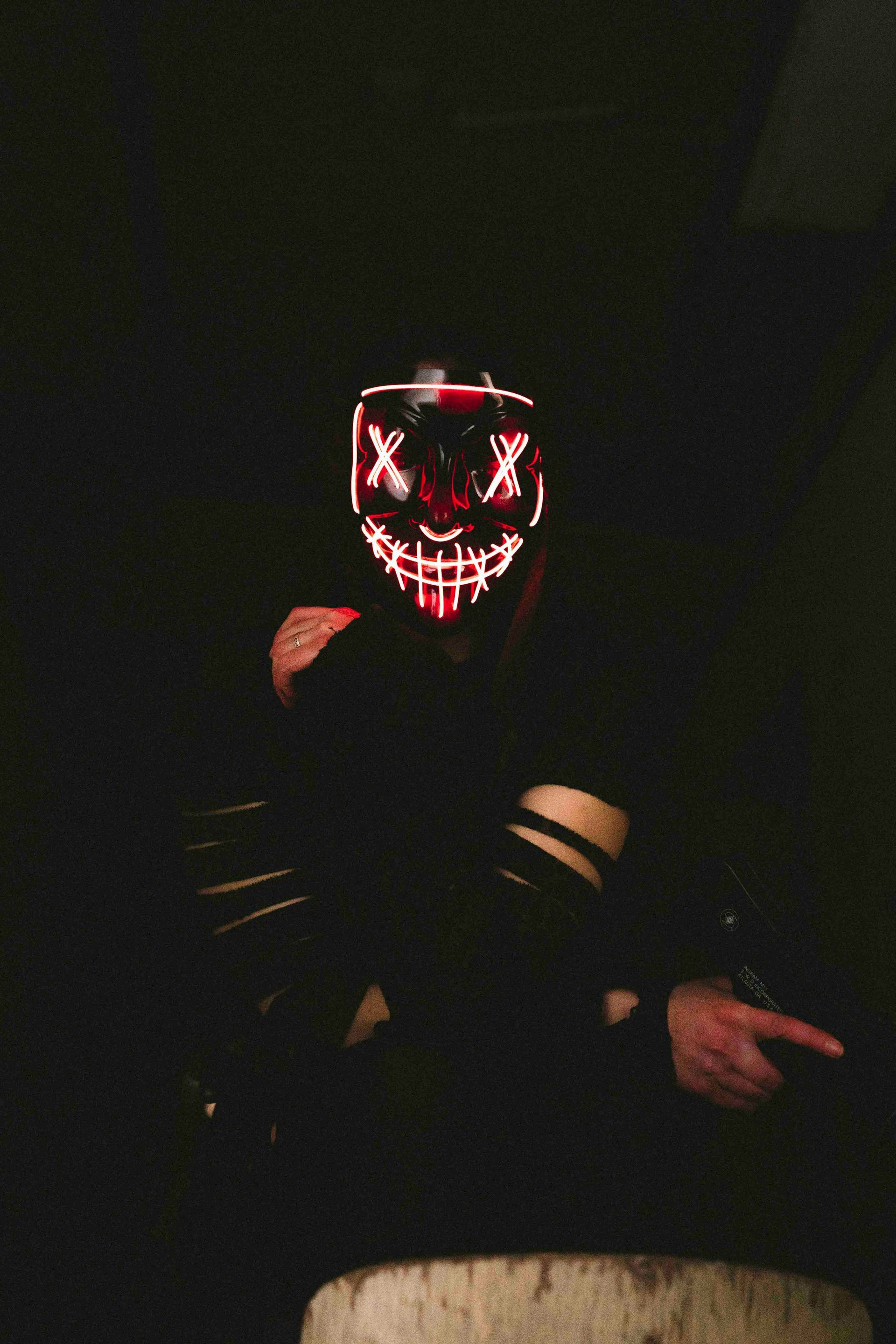 Eine Person mit einer rot leuchtenden „Säuberungs“-Maske zeigt in einem dunklen Raum auf etwas.