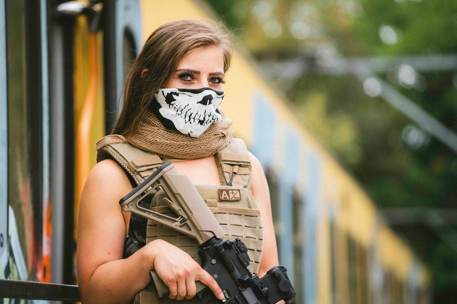 Eine Frau in taktischer Weste und Totenkopfmaske hält ein Gewehr und steht im Freien