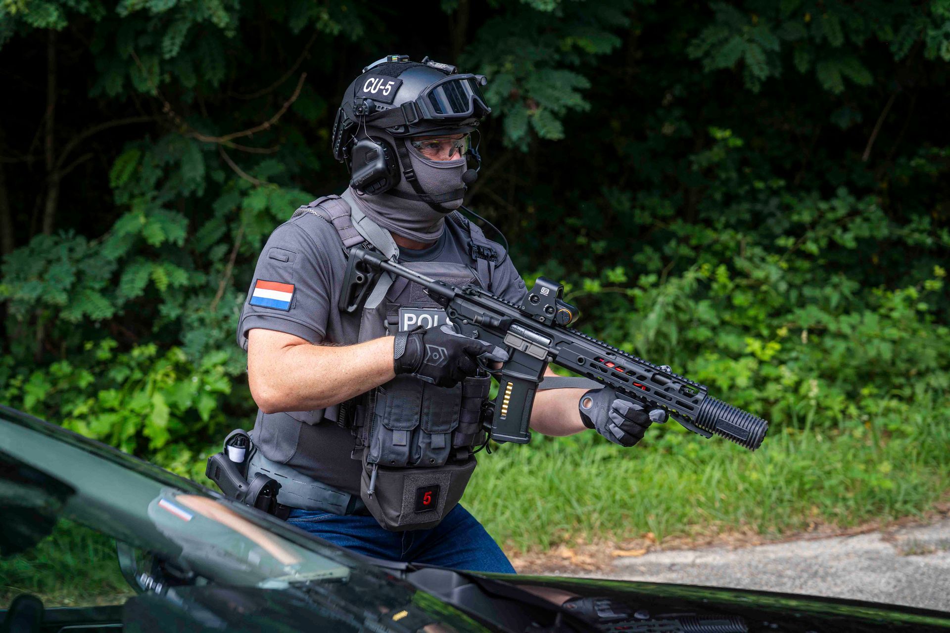 Ein Polizist in taktischer Ausrüstung zielt in einem Waldgebiet mit einem Gewehr in der Nähe eines Fahrzeugs.