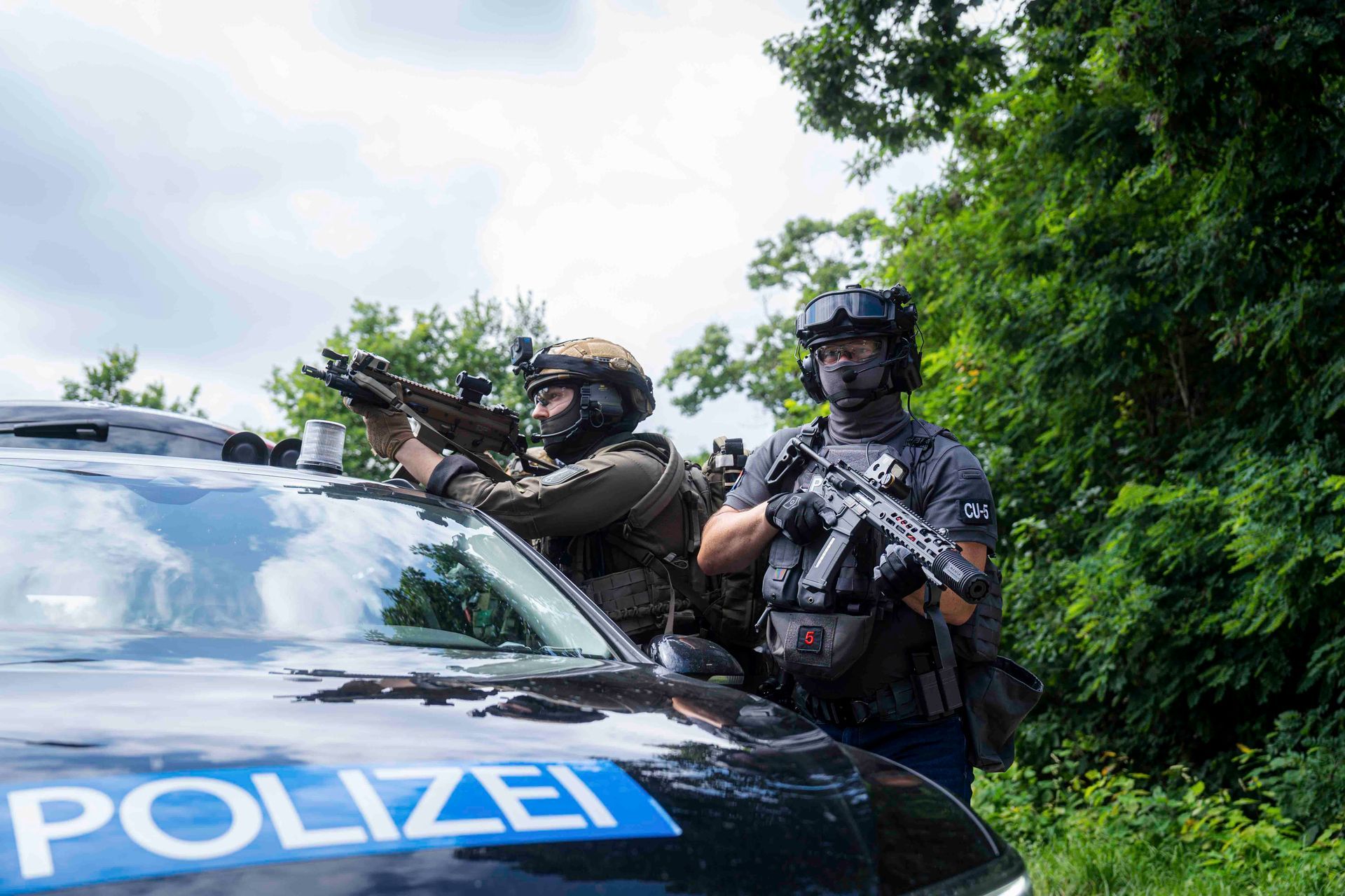 Zwei bewaffnete Polizisten hinter einem schwarz-blauen Polizeiwagen in einem Waldgebiet.