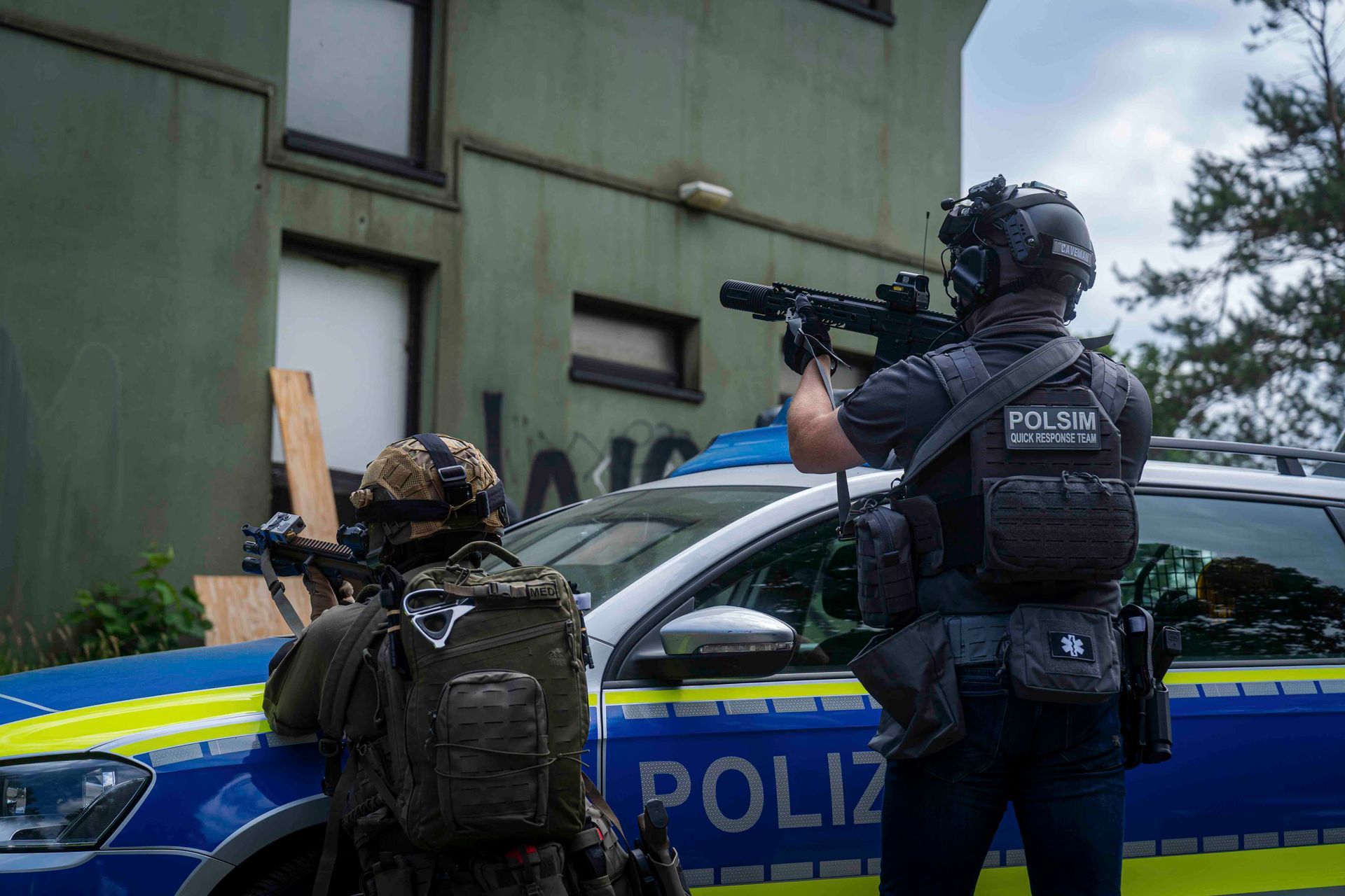 Zwei bewaffnete Polizisten zielen mit Gewehren in der Nähe eines Polizeiwagens vor einem Gebäude.