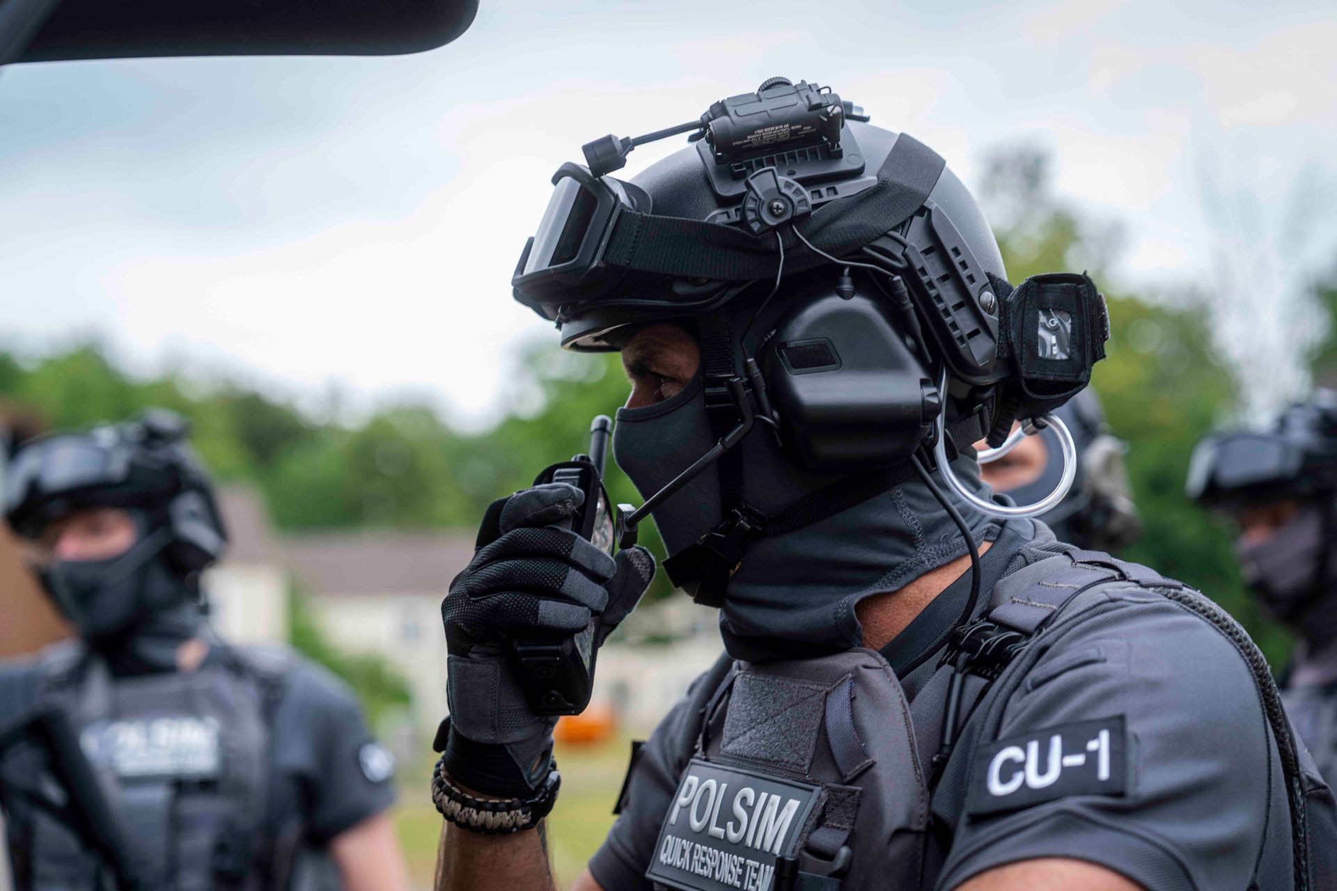 SWAT-Beamter in schwarzer Uniform, Helm mit Headset, Funkgerät, spricht im Freien vor verschwommenem Hintergrund.