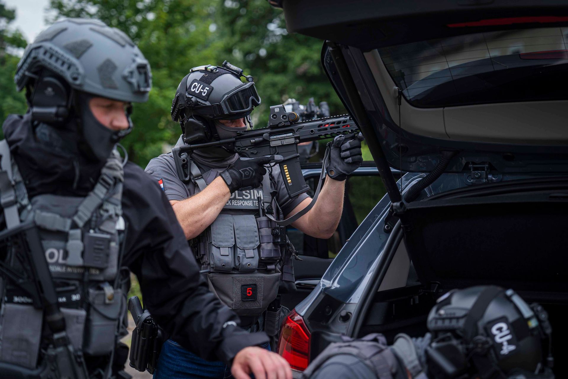 SWAT-Beamte in taktischer Ausrüstung, einer zielt mit einem Gewehr aus einem Fahrzeug, andere Beamte in der Nähe, im Freien.