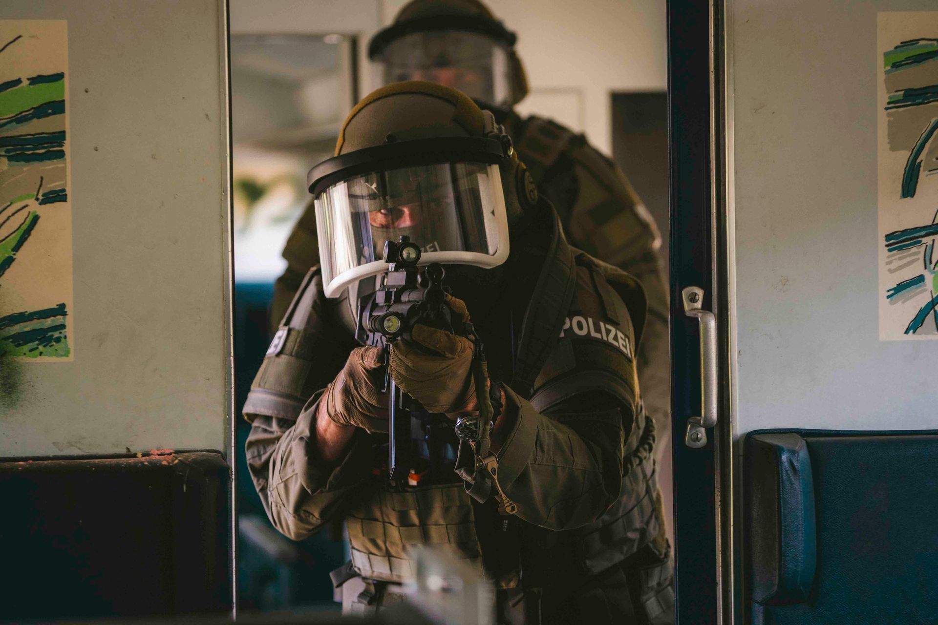 Zwei Polizisten in taktischer Ausrüstung mit Helmen und Waffen in einem Zugwaggon, einer zielt mit einem Gewehr.
