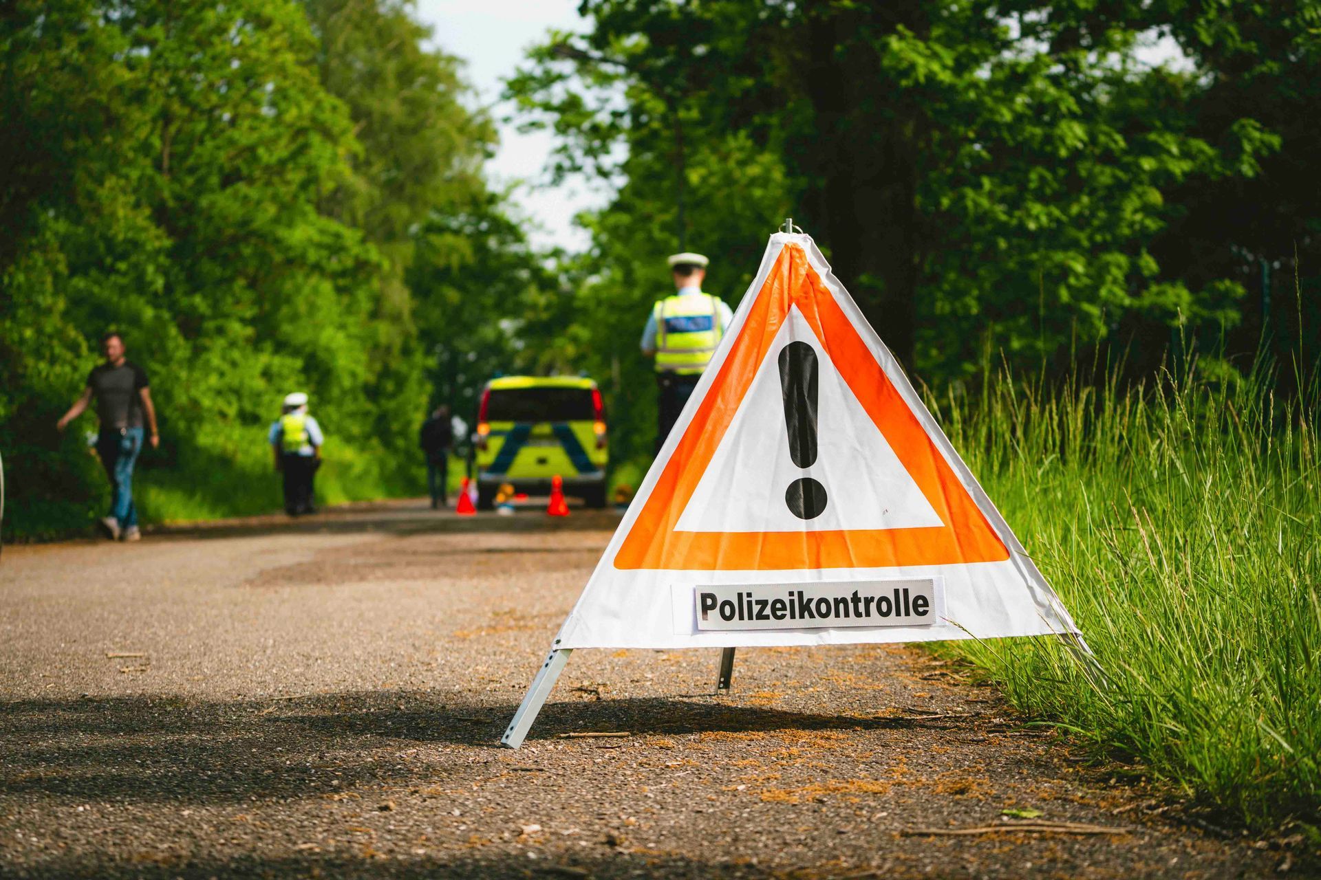 Warnschild mit Ausrufezeichen vor Polizisten und einem Auto auf einer Straße in einem Waldgebiet.