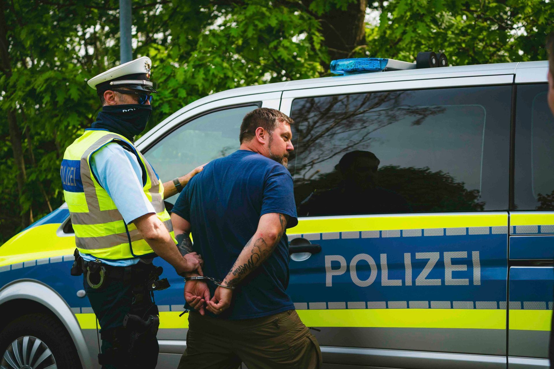 Ein Polizist in einer Warnweste legt einem Mann neben einem Polizeiwagen Handschellen an.