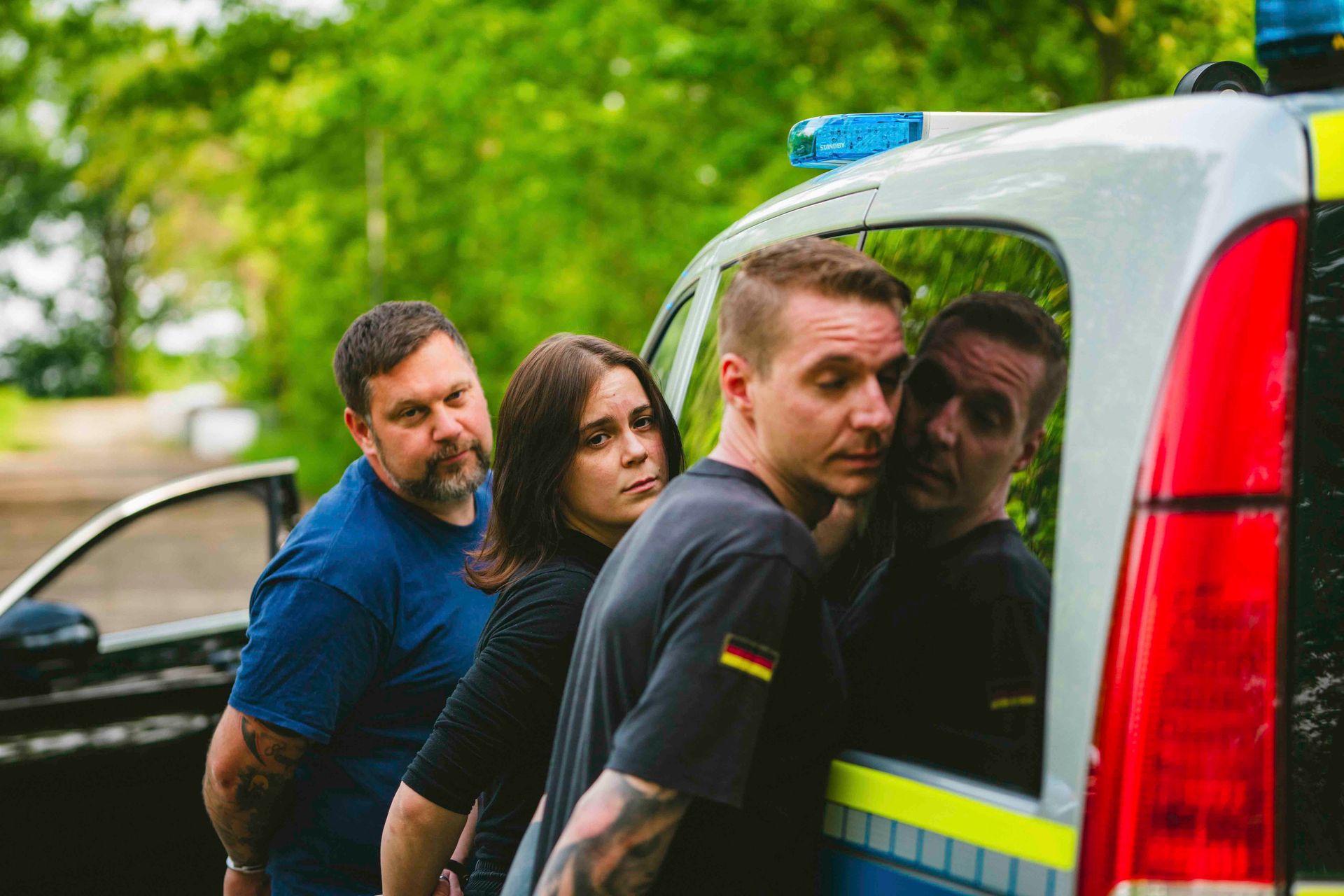Drei Personen mit auf dem Rücken gefesselten Händen neben einem Polizeiwagen, möglicherweise werden sie verhaftet.