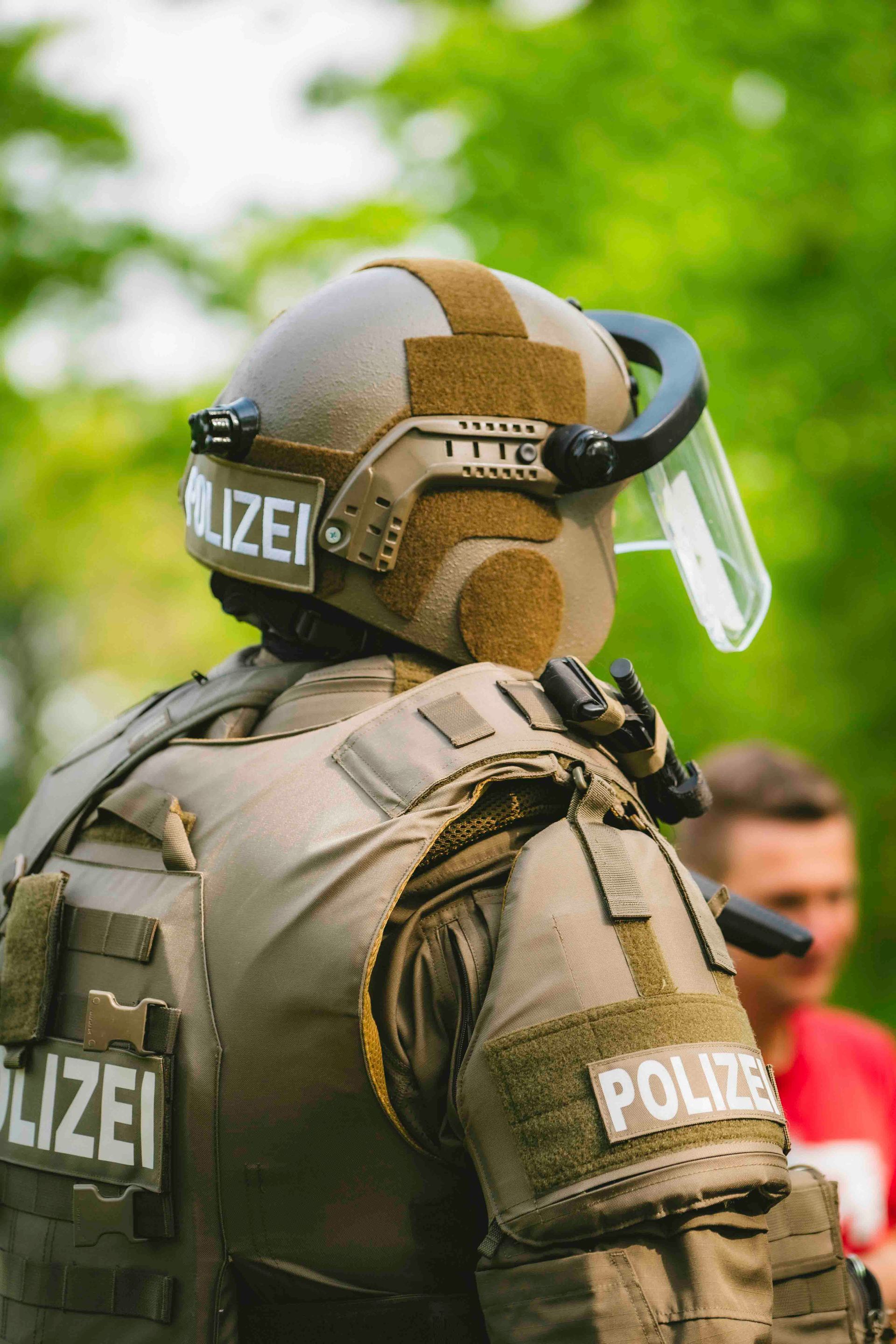 Ein Polizist in taktischer Ausrüstung, Helm mit Visier, steht im Freien. Das Abzeichen „POLIZEI“ ist sichtbar.