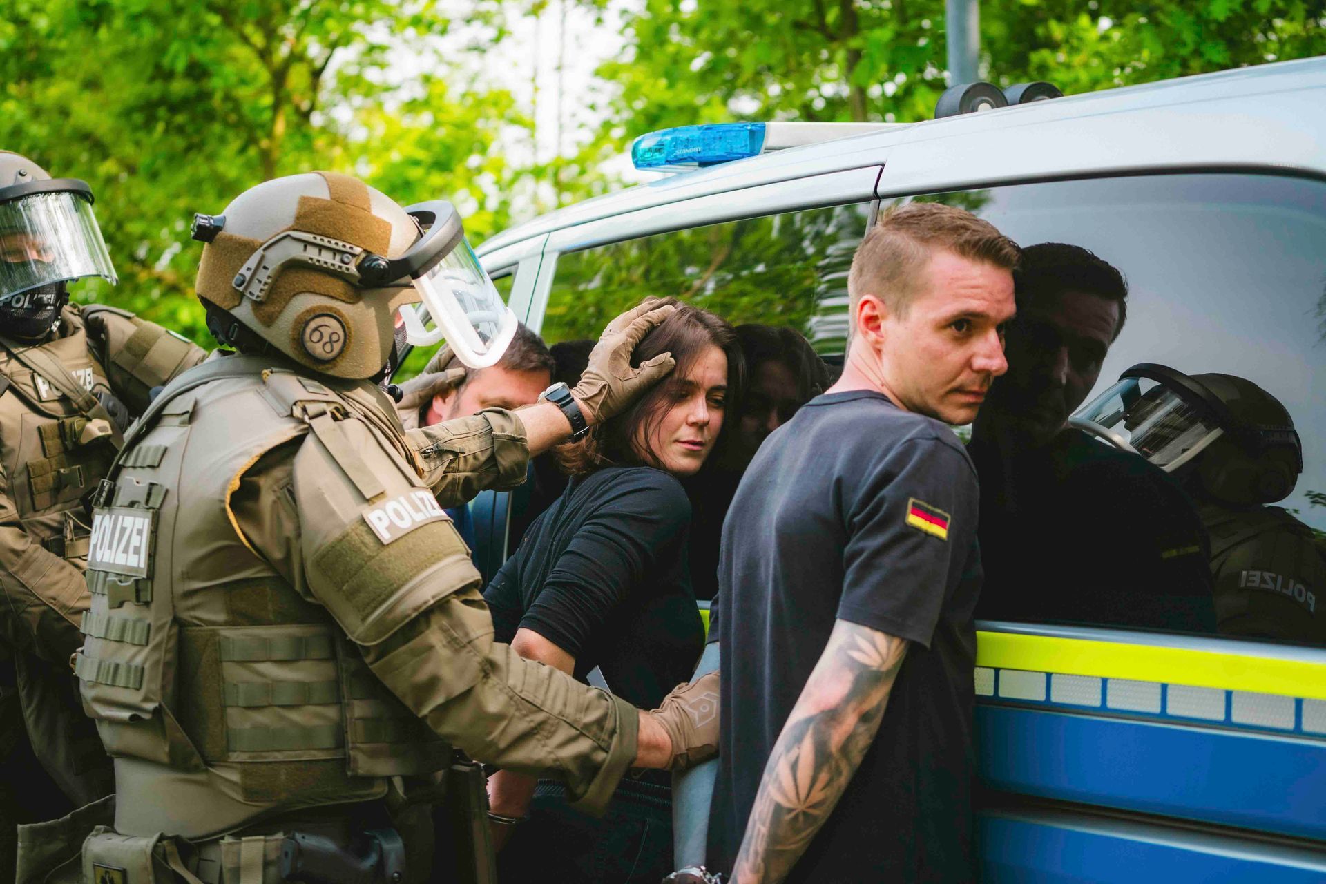 Polizisten nehmen im Freien in der Nähe eines Polizeiwagens eine Frau und einen Mann fest. Die Beamten tragen Ausrüstung.