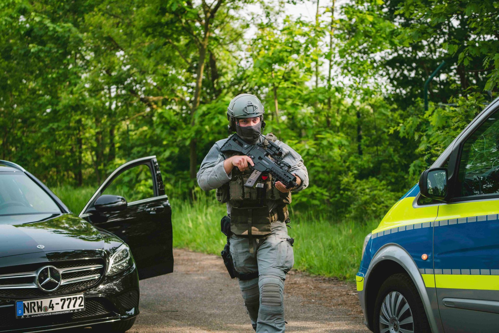 Schwer bewaffneter Polizist steigt in einem Waldgebiet neben einem Polizeiwagen aus einem schwarzen Auto.
