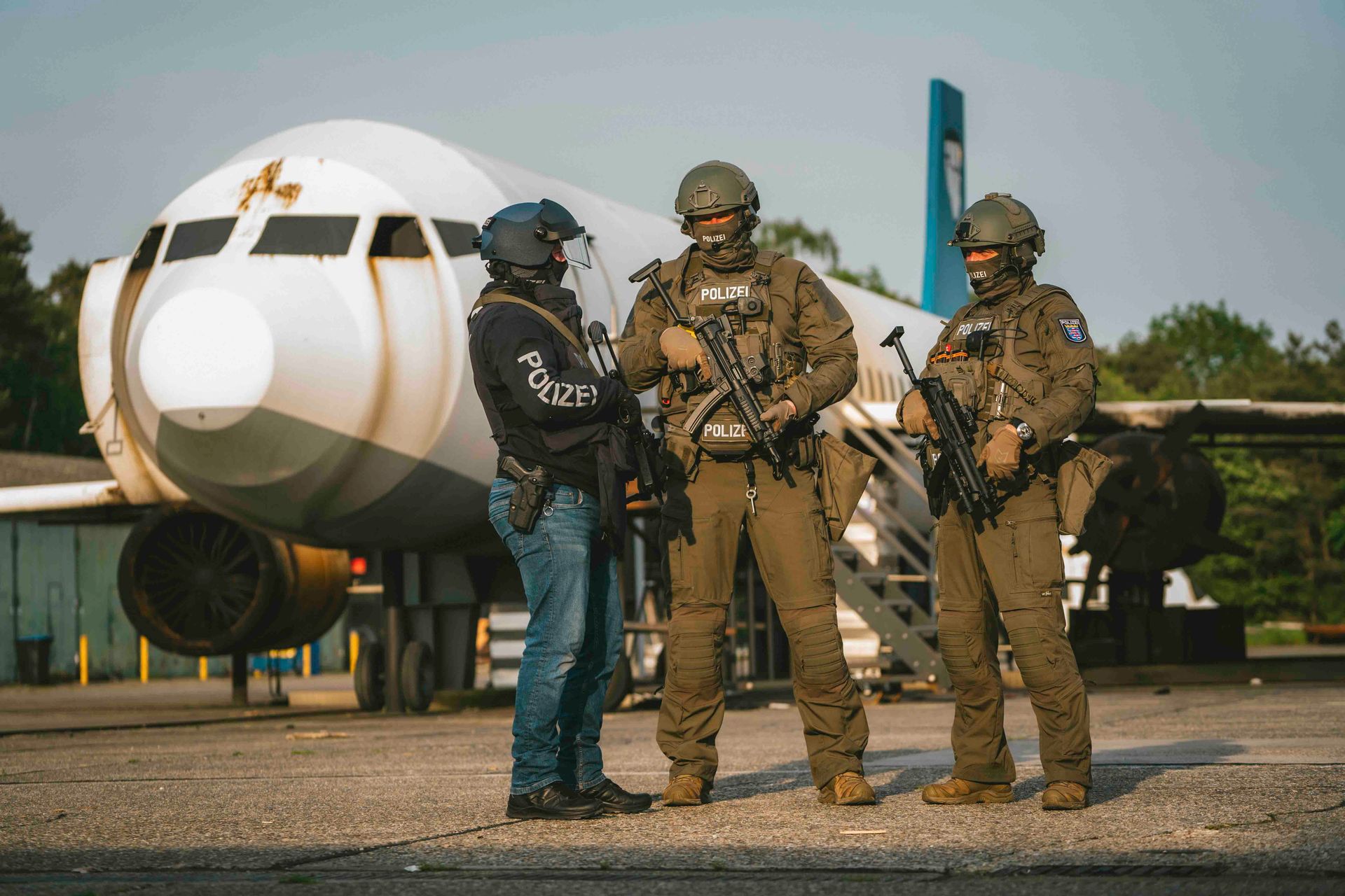 Drei bewaffnete Polizisten und eine Person in Zivilkleidung stehen in der Nähe eines Flugzeugrumpfs.