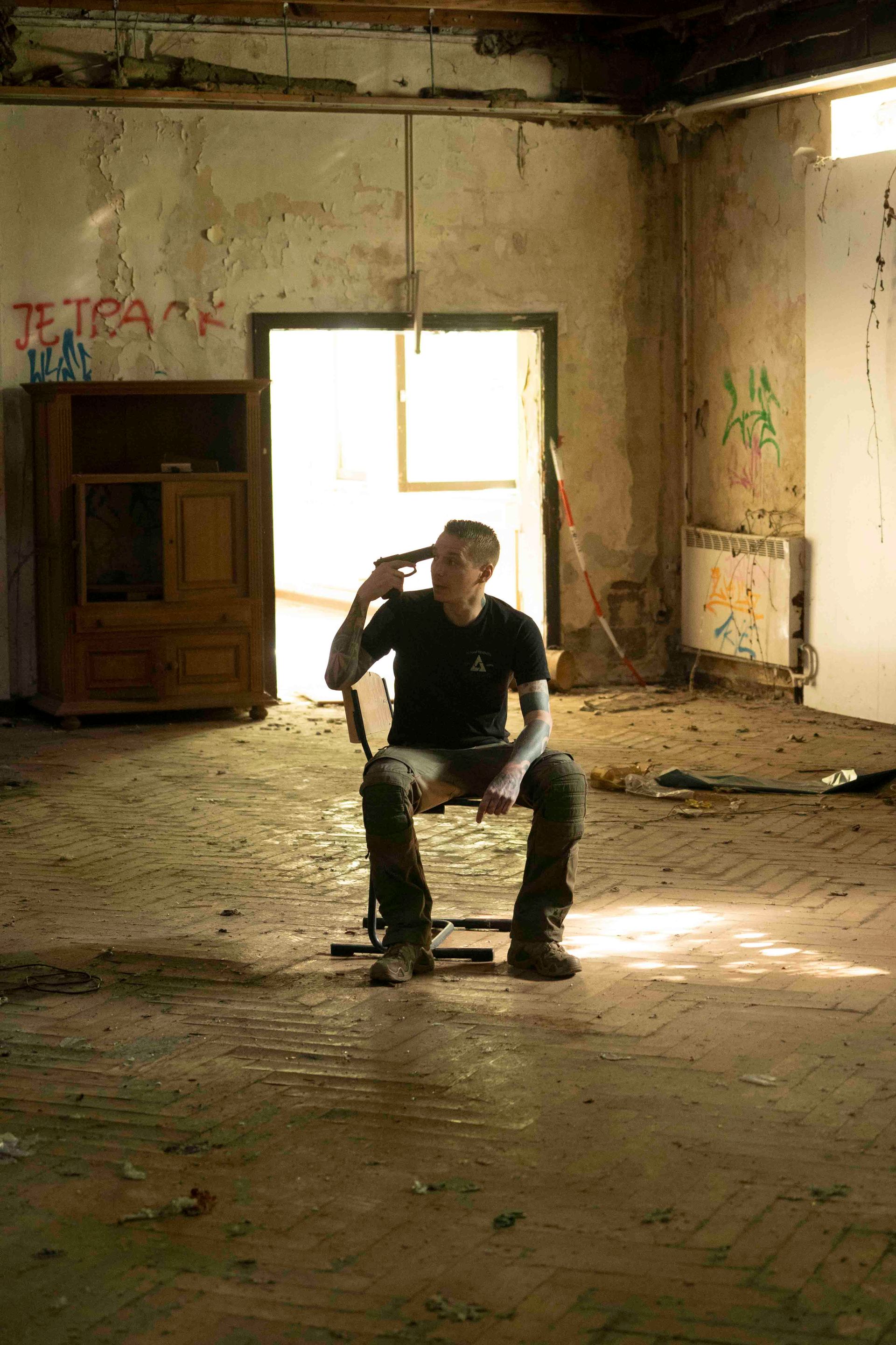 Mann sitzt in einem verlassenen Gebäude auf einem Stuhl und hält sich eine Pistole an den Kopf.