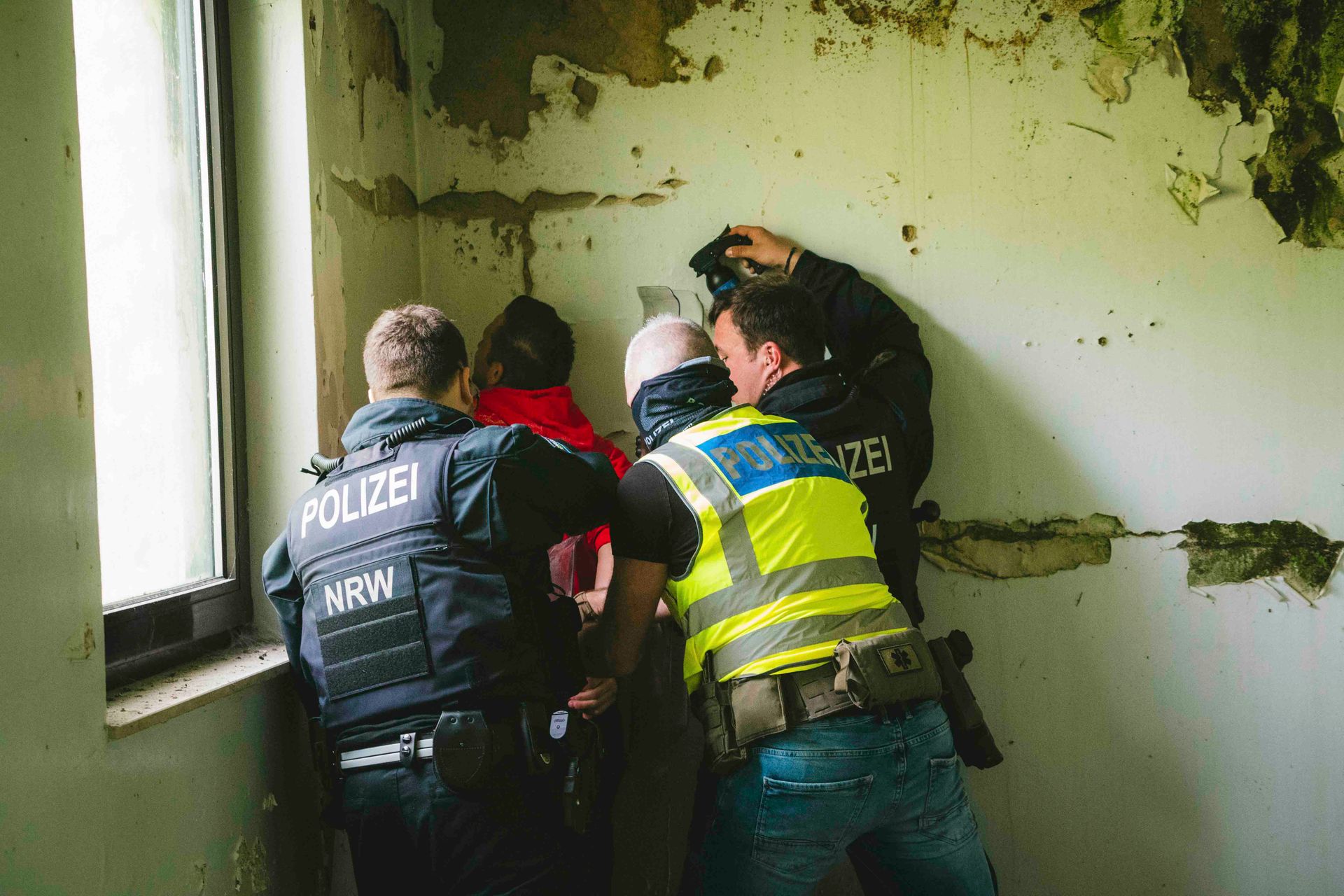 Polizisten drücken eine Person in einem Zimmer gegen eine Wand. Die Wand hat abblätternde Farbe