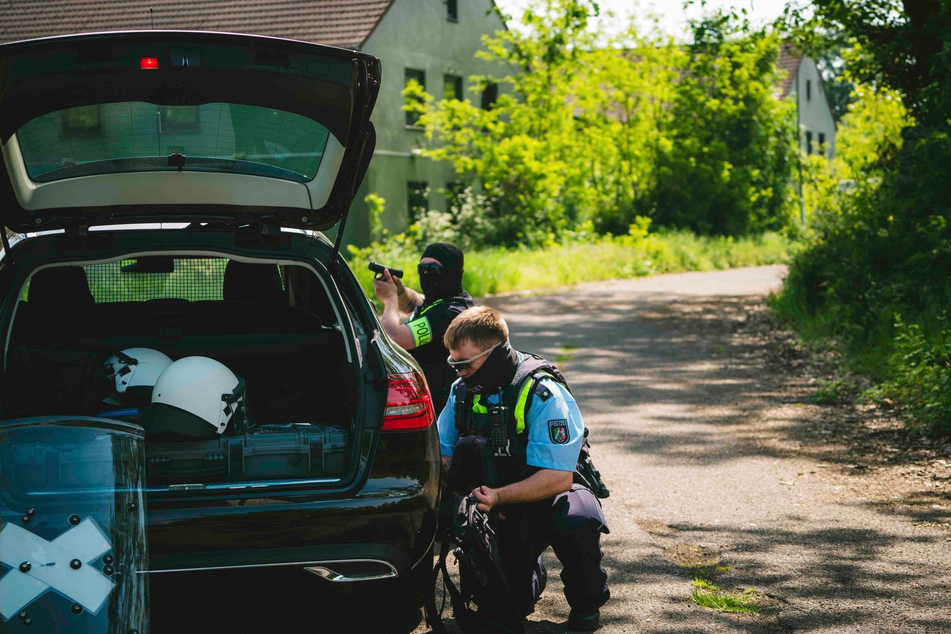 Polizisten stehen neben einem Auto mit geöffnetem Kofferraum; einer zielt mit einer Waffe