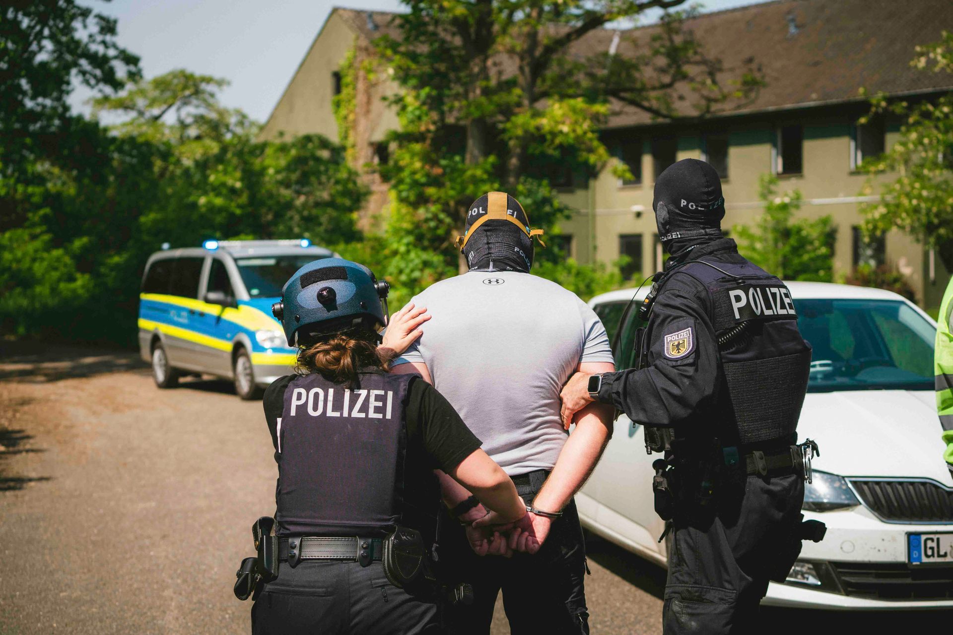 Polizisten nehmen eine Person auf der Straße fest. Im Hintergrund sind ein Polizeiwagen und ein Gebäude zu sehen.