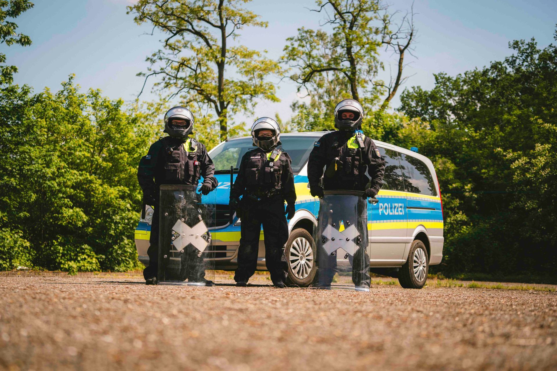 Drei Polizisten in Helmen und Schutzausrüstung stehen vor einem Polizeiwagen in einem Waldgebiet.