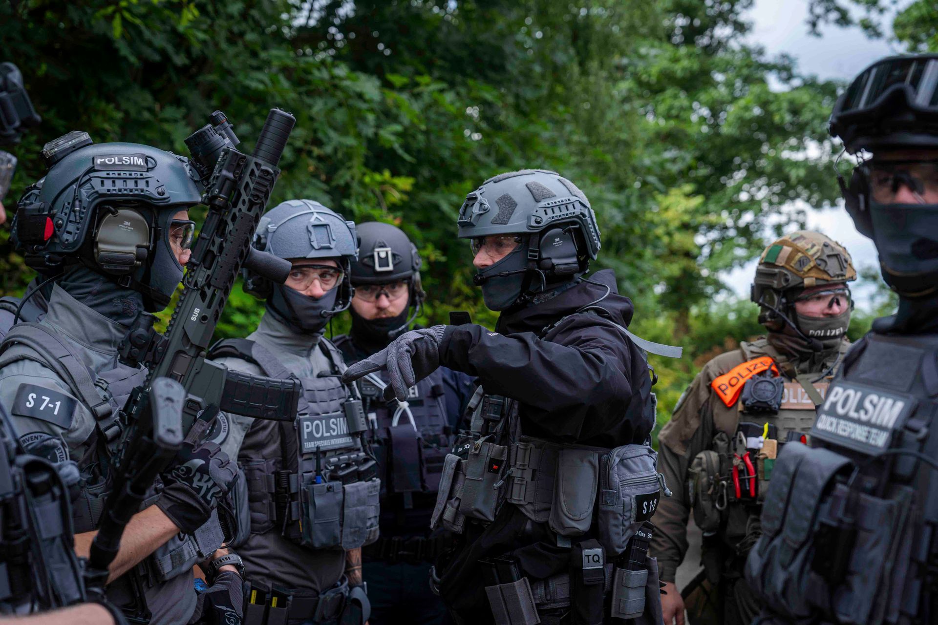Schwer bewaffnete Polizisten in taktischer Ausrüstung, einer zeigt im Freien in der Nähe von Gebüsch.