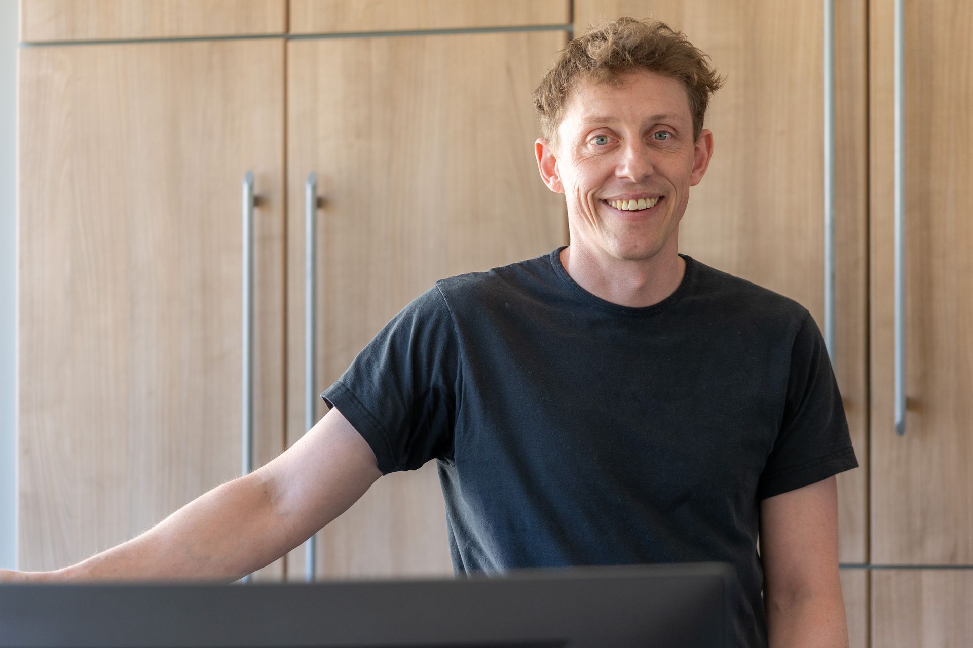 Ein Mann in einem schwarzen Hemd steht vor einem Computermonitor und lächelt.