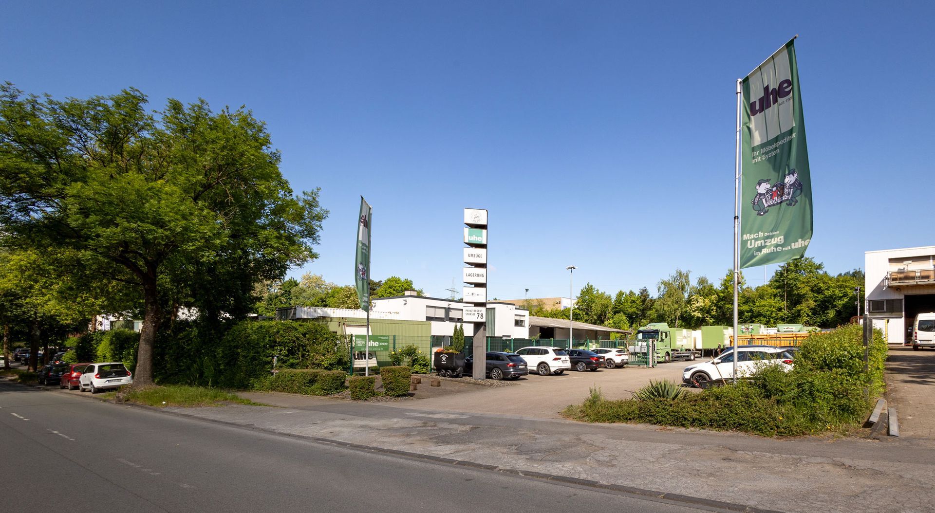 Parkplatz und Geschäftsgebäude unter strahlend blauem Himmel, zwischen Bäumen und einem Straßenschild.