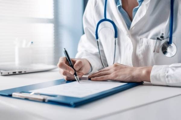 Un médico escribiendo en un portapapeles con un estetoscopio alrededor del cuello en un entorno clínico luminoso.