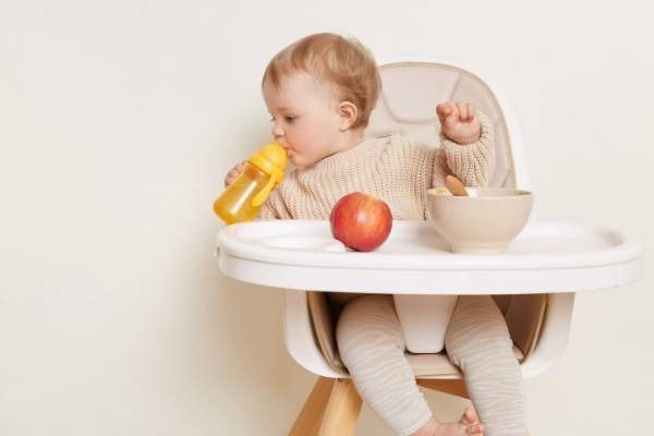 Un niño pequeño sentado en una trona bebe de un biberón amarillo junto a una manzana y un cuenco.