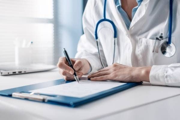 Un médico escribe en un portapapeles en un escritorio, vestido con una bata blanca y con un estetoscopio, en una oficina luminosa.