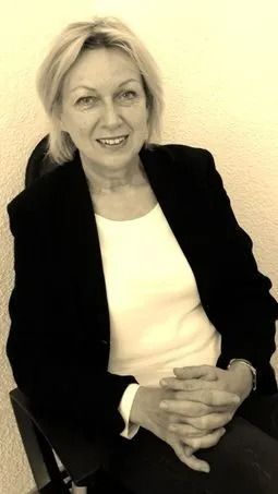 Eine lächelnde Frau in einem schwarzen Blazer und einem weißen Top sitzt mit verschränkten Händen an einer hellen Wand.