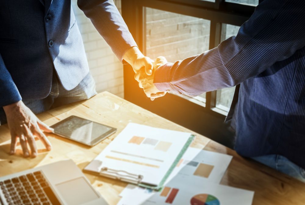 Dos personas con traje se dan la mano sobre un escritorio con documentos y un ordenador portátil.