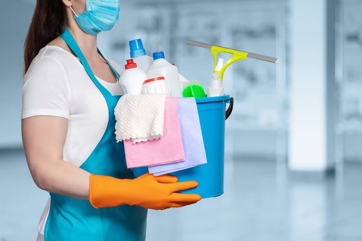 Una mujer que lleva una máscara y guantes sostiene un balde con productos de limpieza.