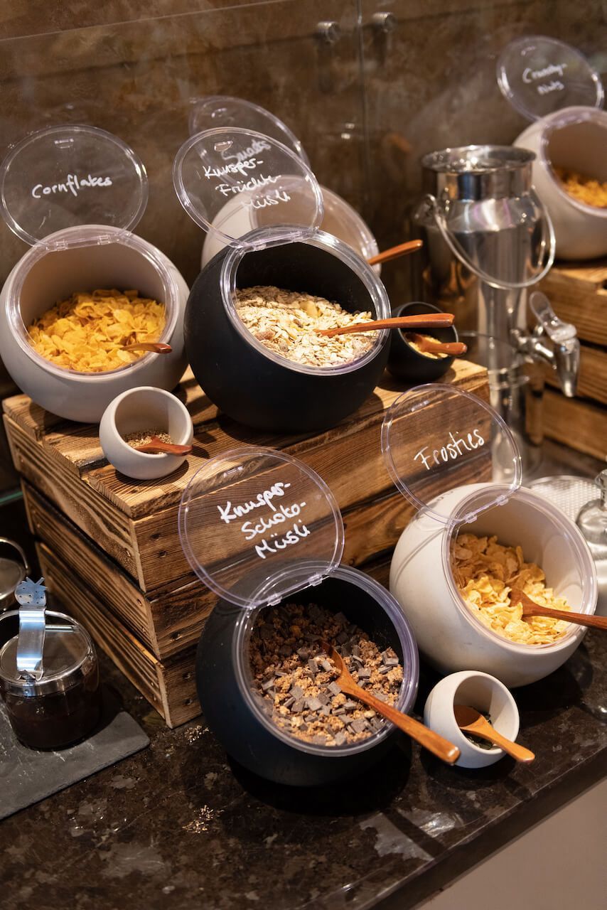 Buffet-Müslistation mit verschiedenen Müslisorten in schwarzen Schüsseln, Schaufeln und Glasspendern auf einem Holzgestell.