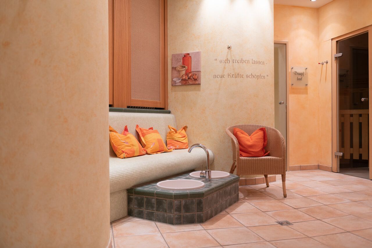 Gemütliche Wellness-Oase mit orangefarbenen Kissen, Korbsessel, Fliesenboden und warmen beigefarbenen Wänden.