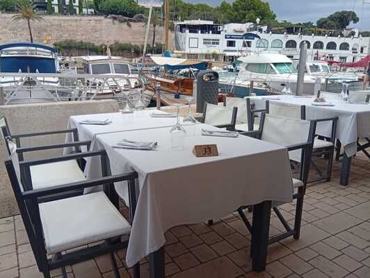 Mesas de restaurante al aire libre dispuestas con manteles y sillas blancas, con vistas a un puerto deportivo con barcos.