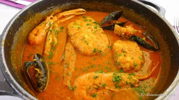 Guiso de mariscos con camarones, pescado y mejillones en un caldo de color naranja rojizo, adornado con perejil, en una olla de metal.