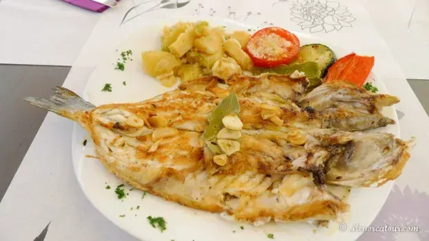 Pescado a la parrilla, patatas, tomate y calabacín en un plato blanco, posiblemente en un restaurante.
