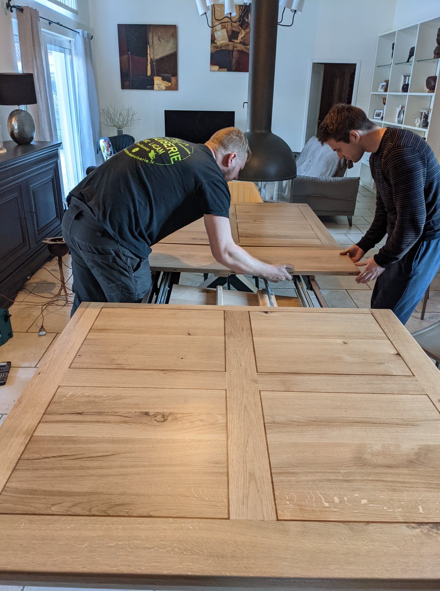 Deux personnes assemblent une grande table en bois dans un atelier lumineux, avec un poêle noir en arrière-plan.